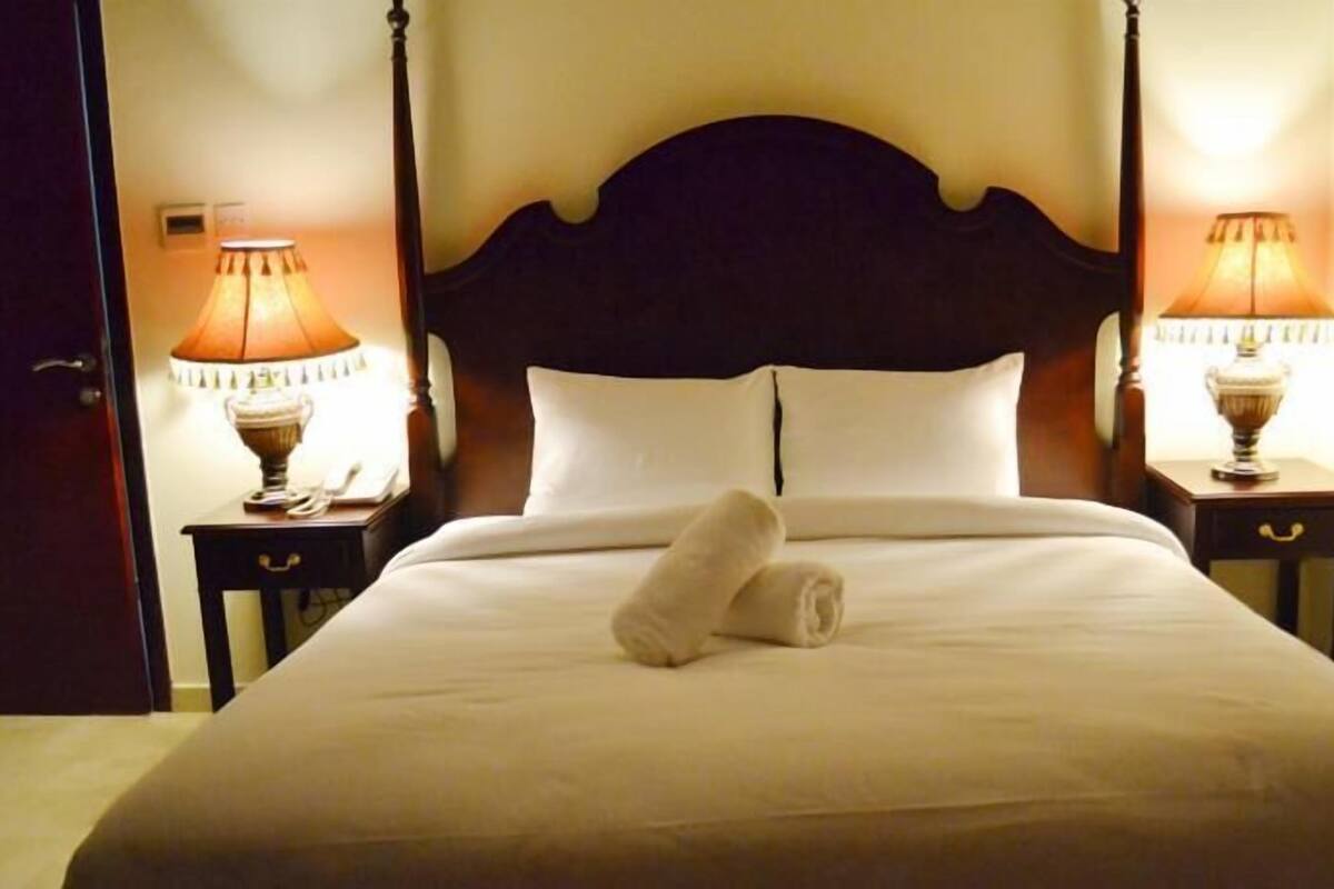 A neatly made bed with a rolled towel, two bedside lamps, and a dark wooden headboard.