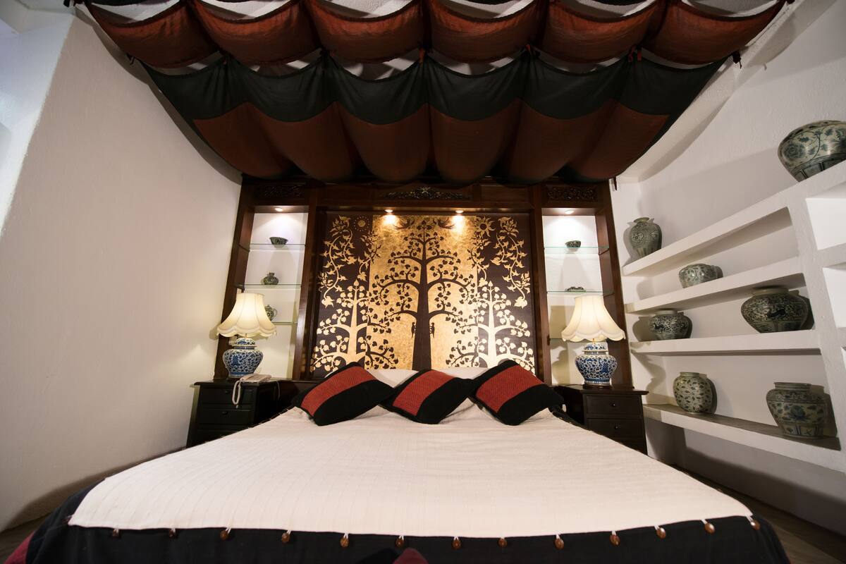 Une chambre à coucher bien aménagée, avec un lit, deux tables de chevet dotées de lampes, un panneau décoratif au mur et des étagères ornées de vases.