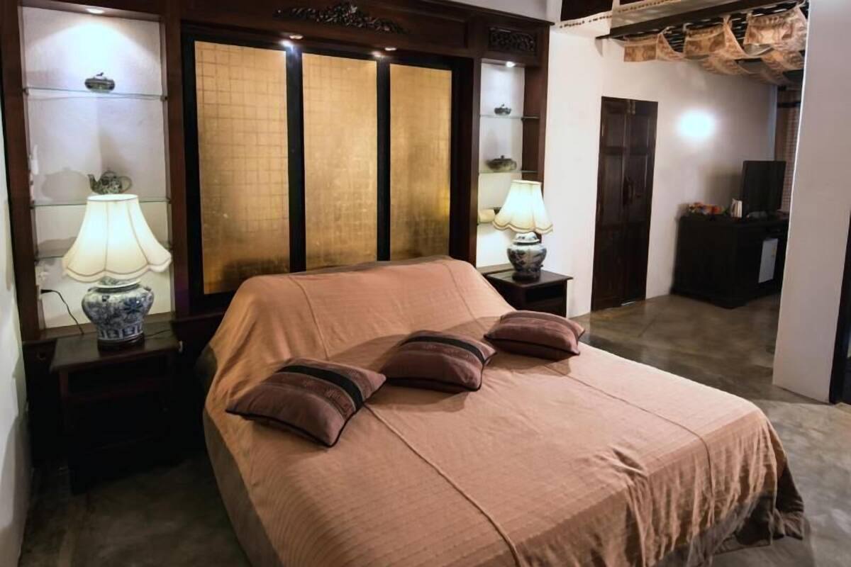 Un lit bien fait, recouvert d’une couverture marron et agrémenté de trois oreillers, avec deux tables de chevet de chaque côté, sur lesquelles sont posées des lampes.