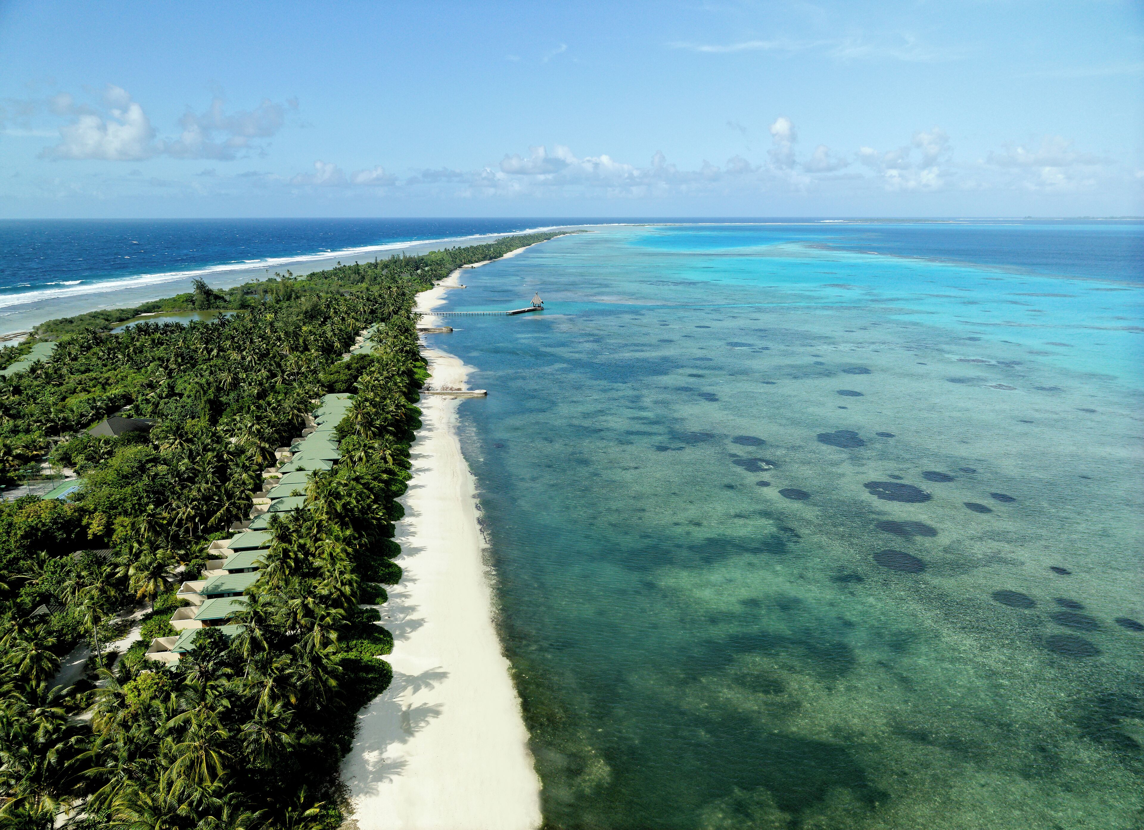 An aerial view of a beach with clear turquoise waters, a sandy shoreline, and dense tropical vegetation.