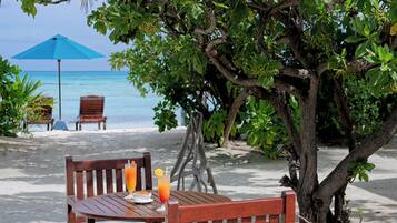 A wooden table with two chairs and a blue umbrella, overlooking a beach with clear blue water.