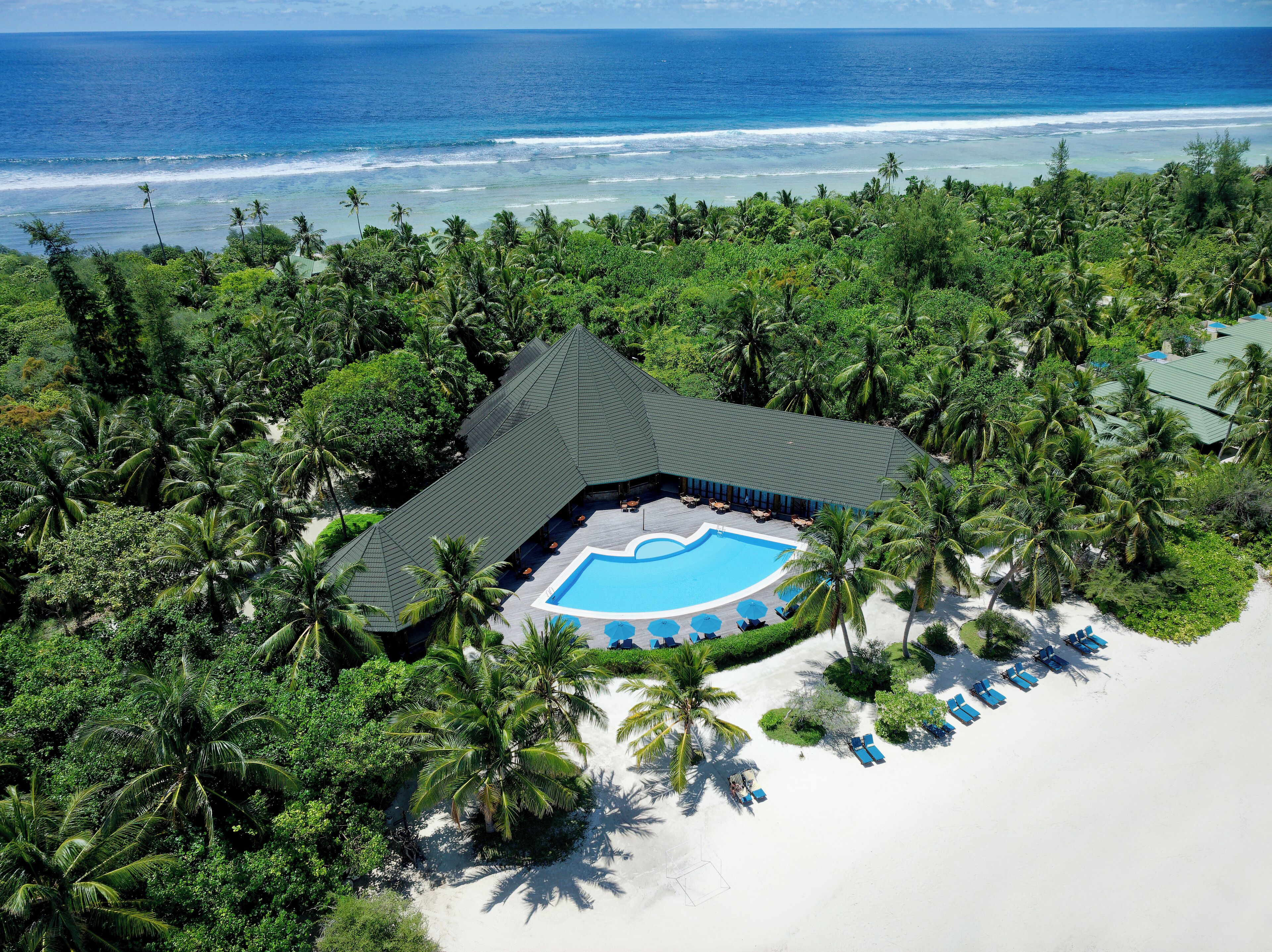 Eine Vogelperspektive zeigt ein Resort mit einem Schwimmbad, das von Palmen und einem Strand umgeben ist.