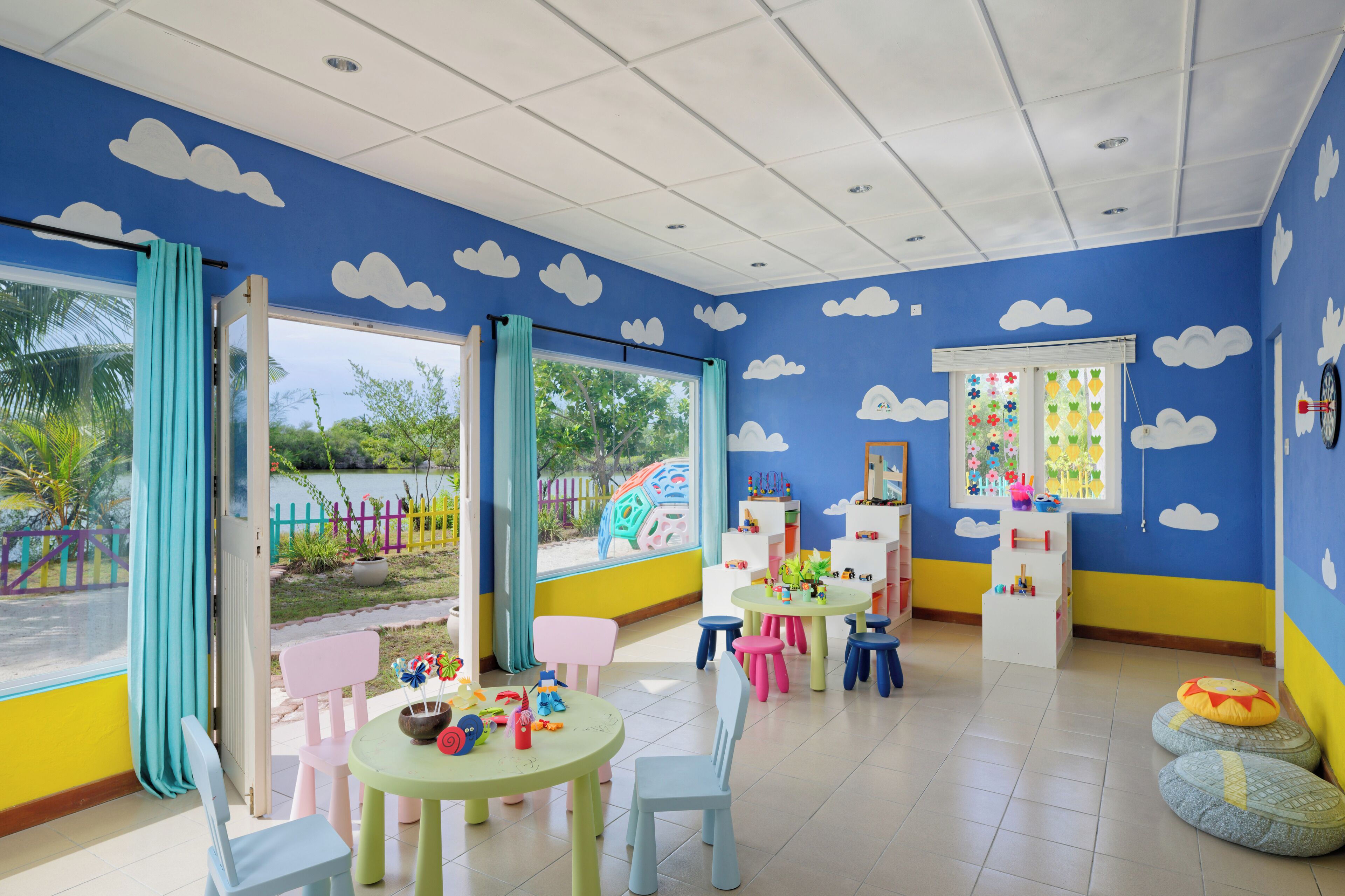 Une salle de jeux pour enfants avec des meubles aux couleurs vives, une vue sur un terrain de jeux et un mur bleu décoré de motifs de nuages.