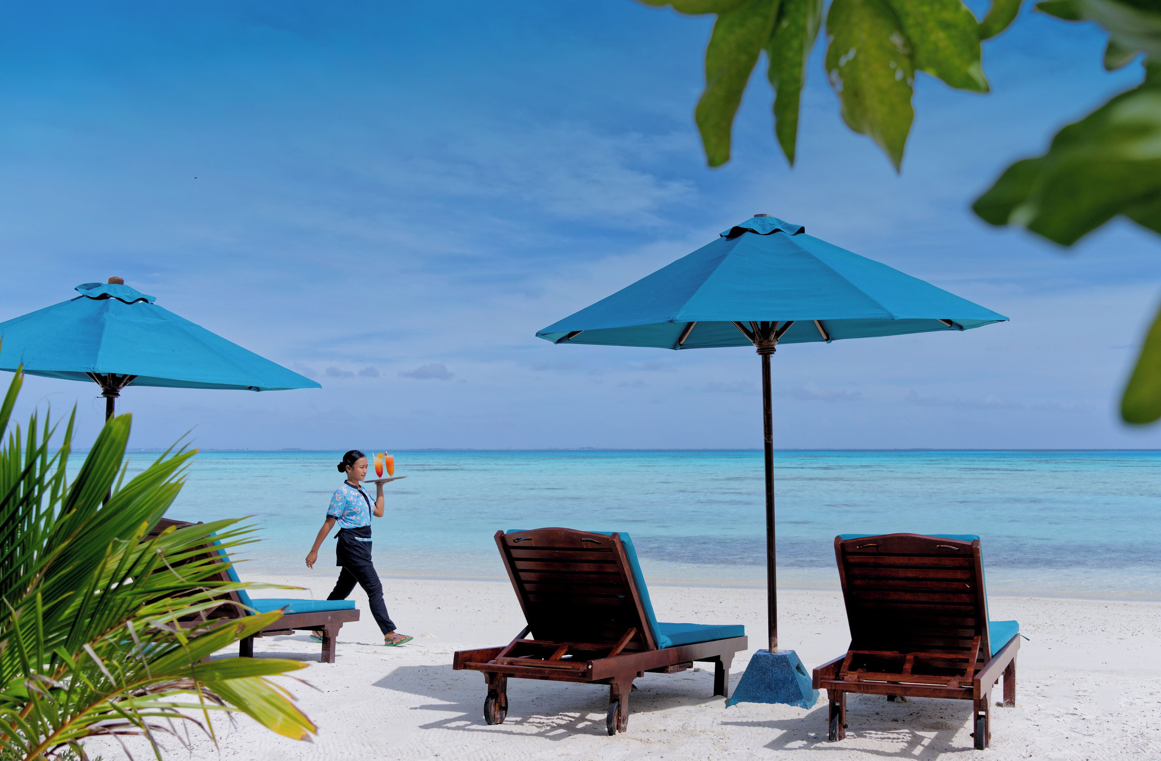 A beach with two wooden lounge chairs and two blue umbrellas, a person walking, and a clear view of the ocean.