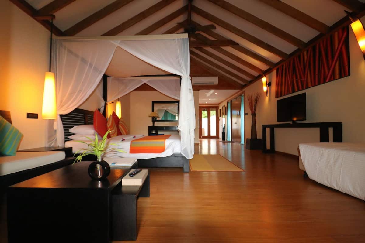 A room with a bed, a sofa, a TV, and a desk with a vase and remote control.