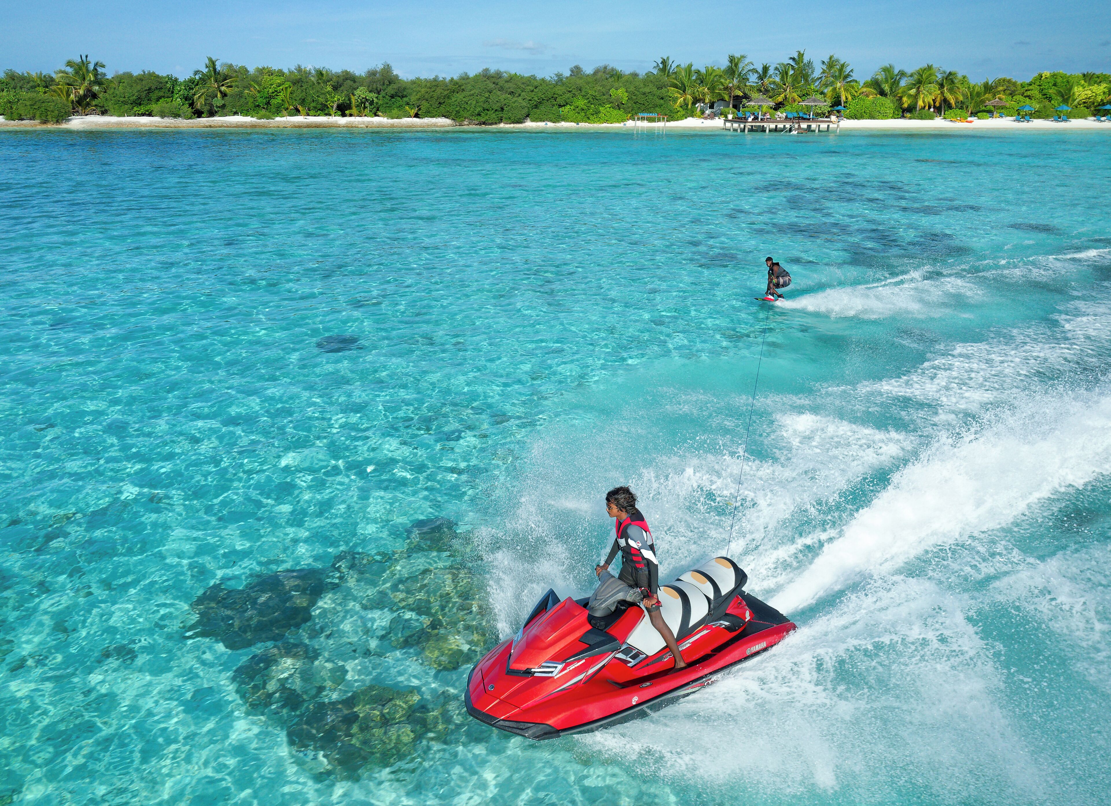 A clear blue sea with a jet ski in the foreground and a person wakeboarding in the background.