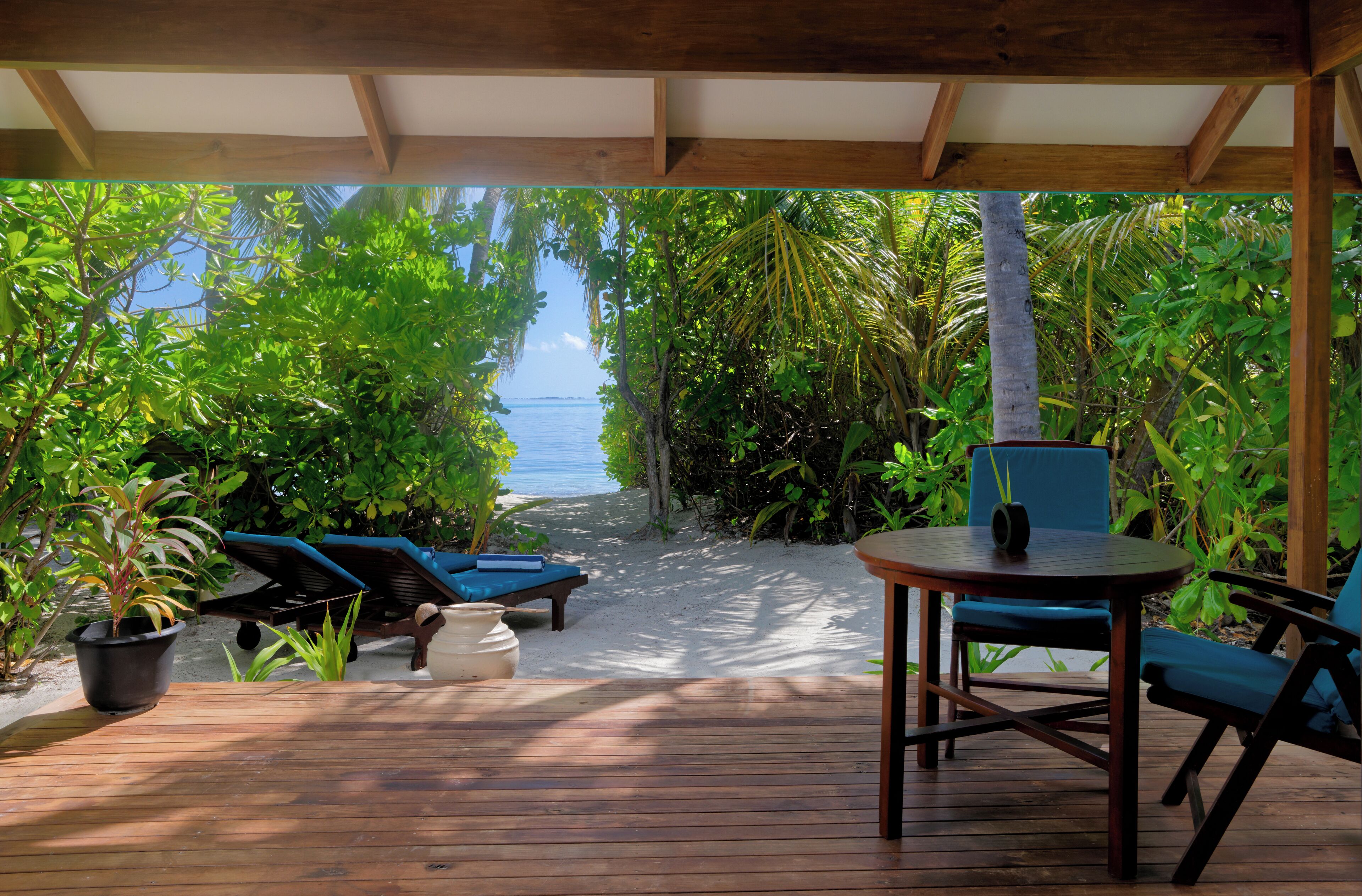 Terraza de madera con sillones y una mesa, rodeada de exuberante vegetación tropical, que da a una playa.