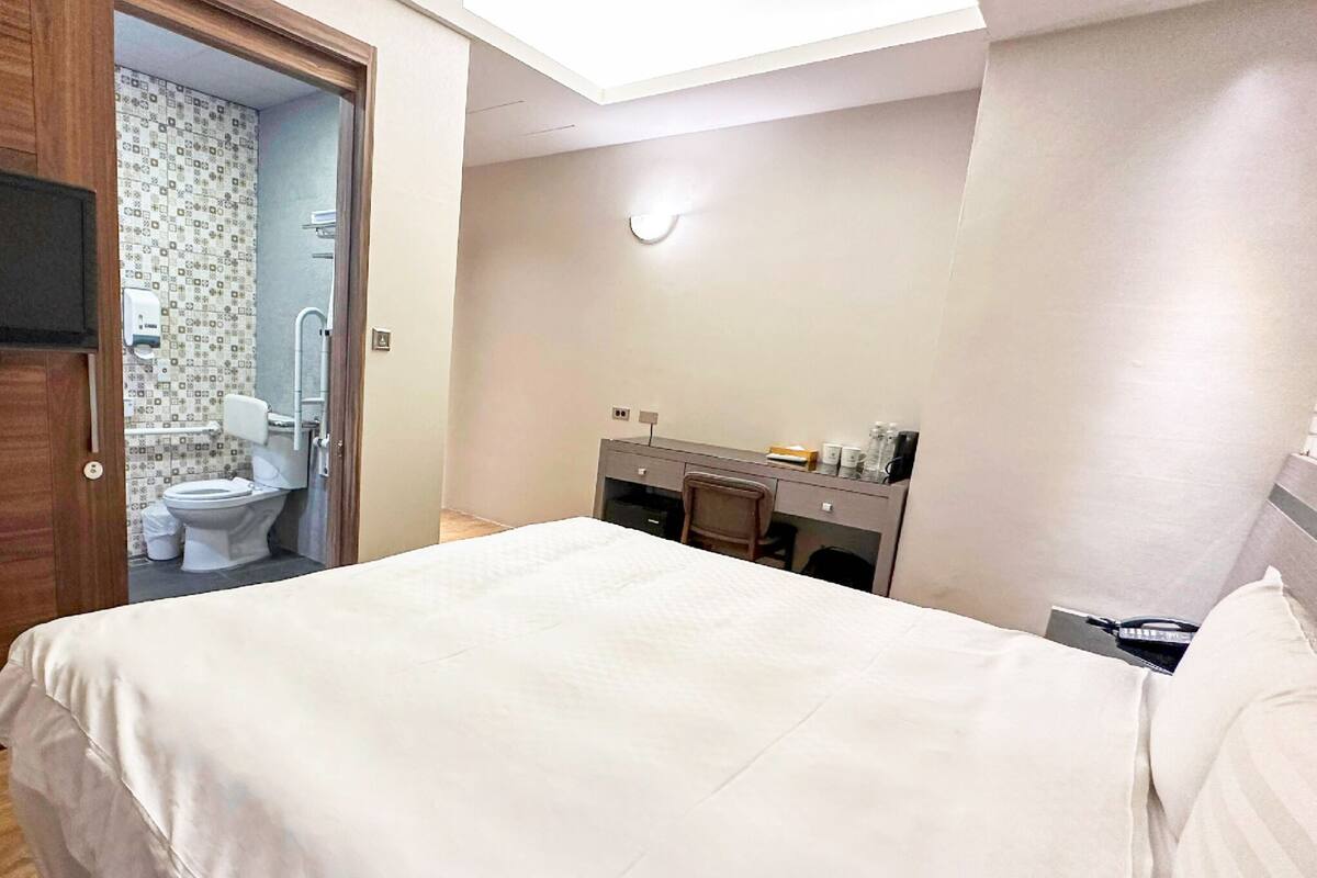 Hotelkamer met een bed, bureau, stoel, televisie, spiegel en een zichtbare badkamer door een open deur.