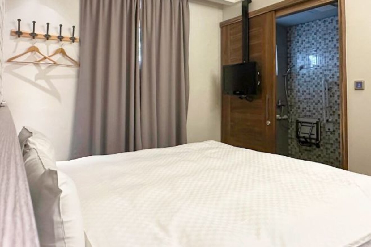 A hotel room with a bed, a wooden wardrobe, a TV mounted on the wardrobe, and a shower area visible through a glass door.