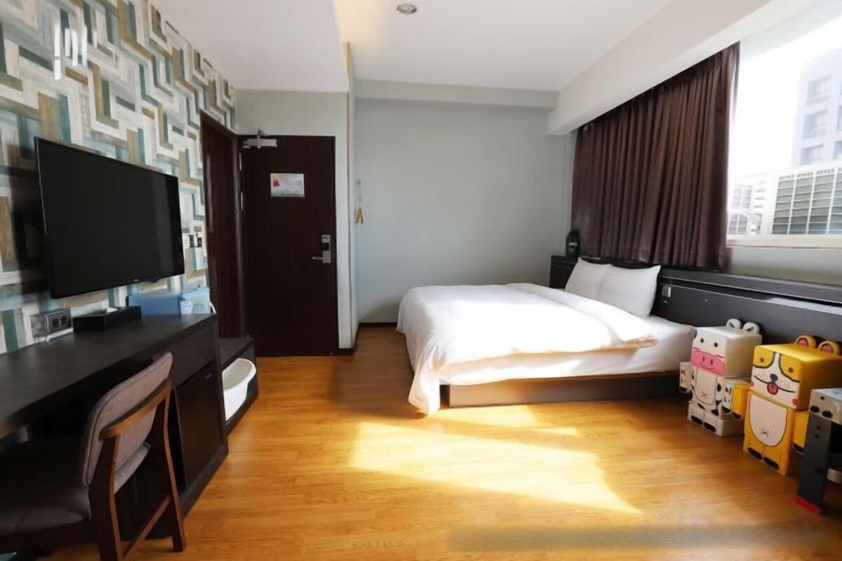 A hotel room with a bed, a desk with a television, a window with curtains, and a wall with a geometric pattern.