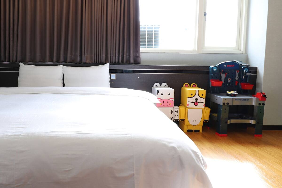 Hotelkamer met een groot bed, een raam met gordijnen, een bureau met een printer en een kleintje tafeltje met speelgoed.