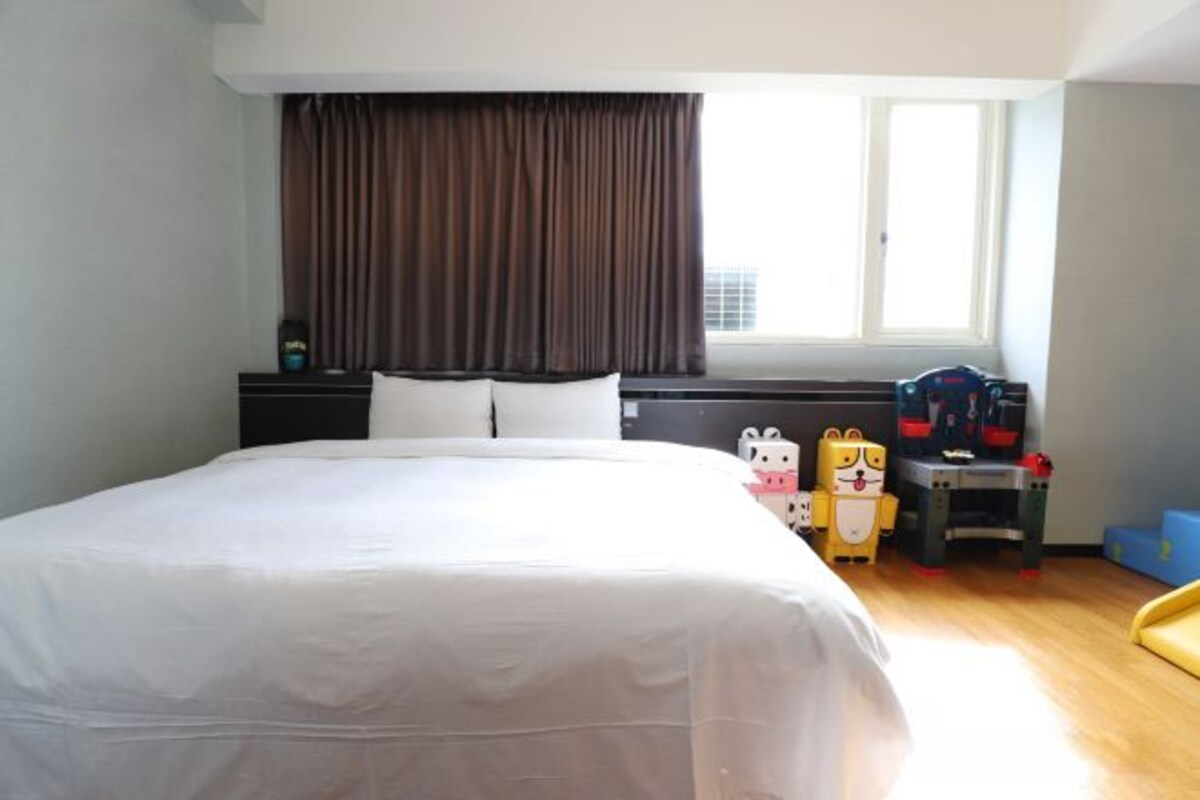 A hotel room with a large bed, a window with curtains, a desk with a chair, and a toy figure on the desk.