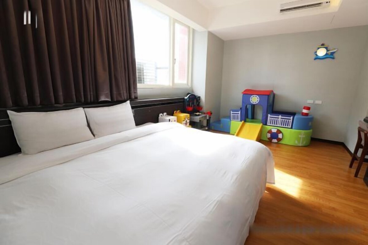A hotel room with a large bed, a window with curtains, a children's play area with toys, and a clock on the wall.