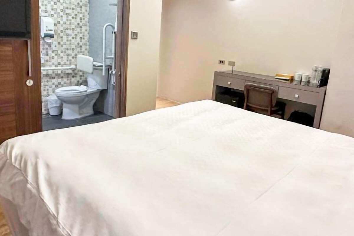 Quarto de hotel com cama, escrivaninha e banheiro visível por uma porta aberta.