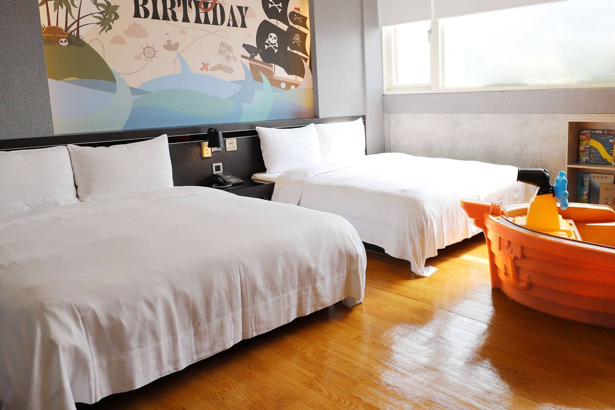 Een hotelkamer met twee bedden, een kinderspeelruimte, een boekenkast en een groot raam.