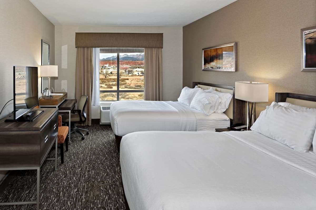 A hotel room with two beds, a desk, a chair, a lamp, and a window with a mountain view.