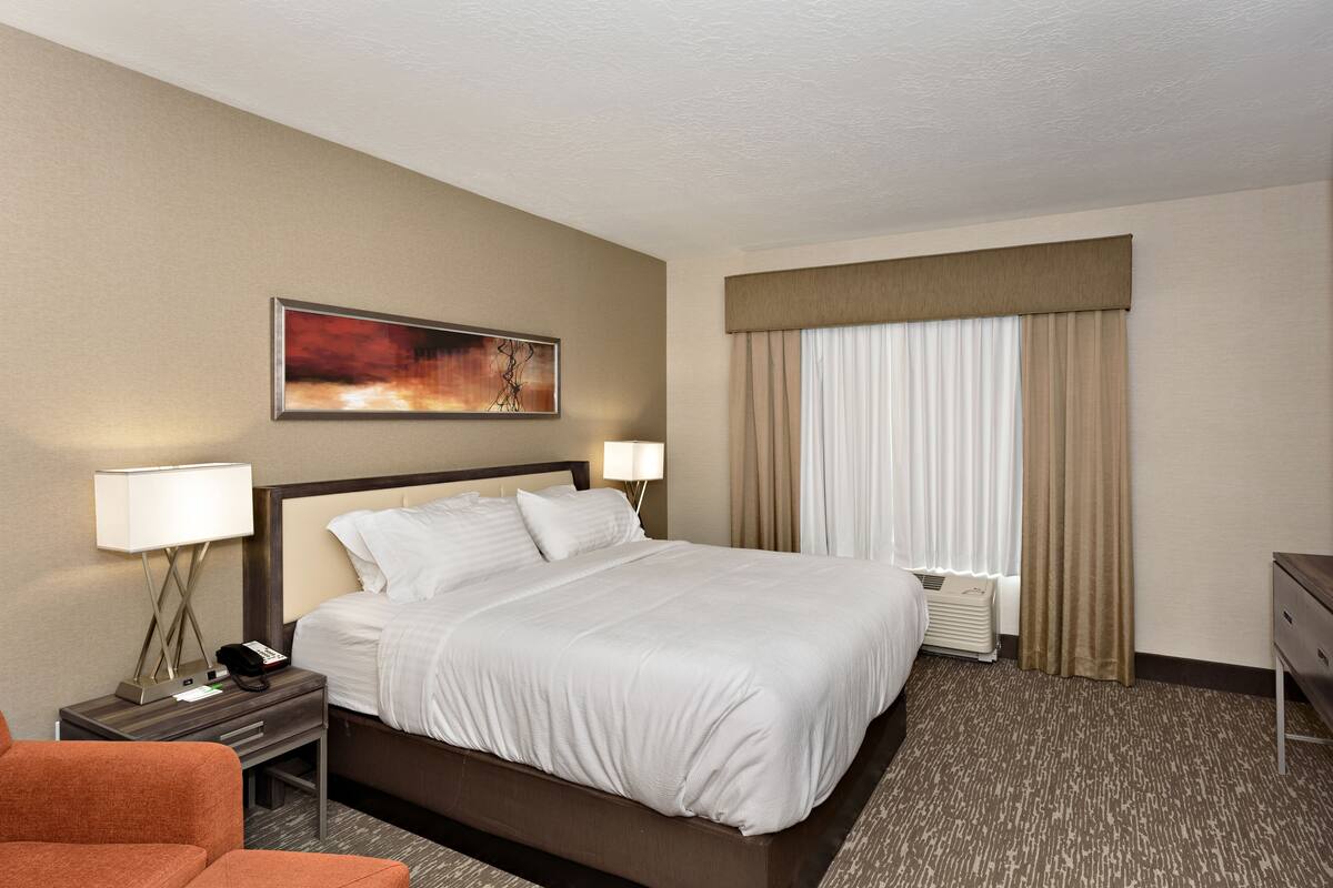 A hotel room with a large bed, two bedside tables with lamps, a chair, a painting on the wall, and a window with curtains.