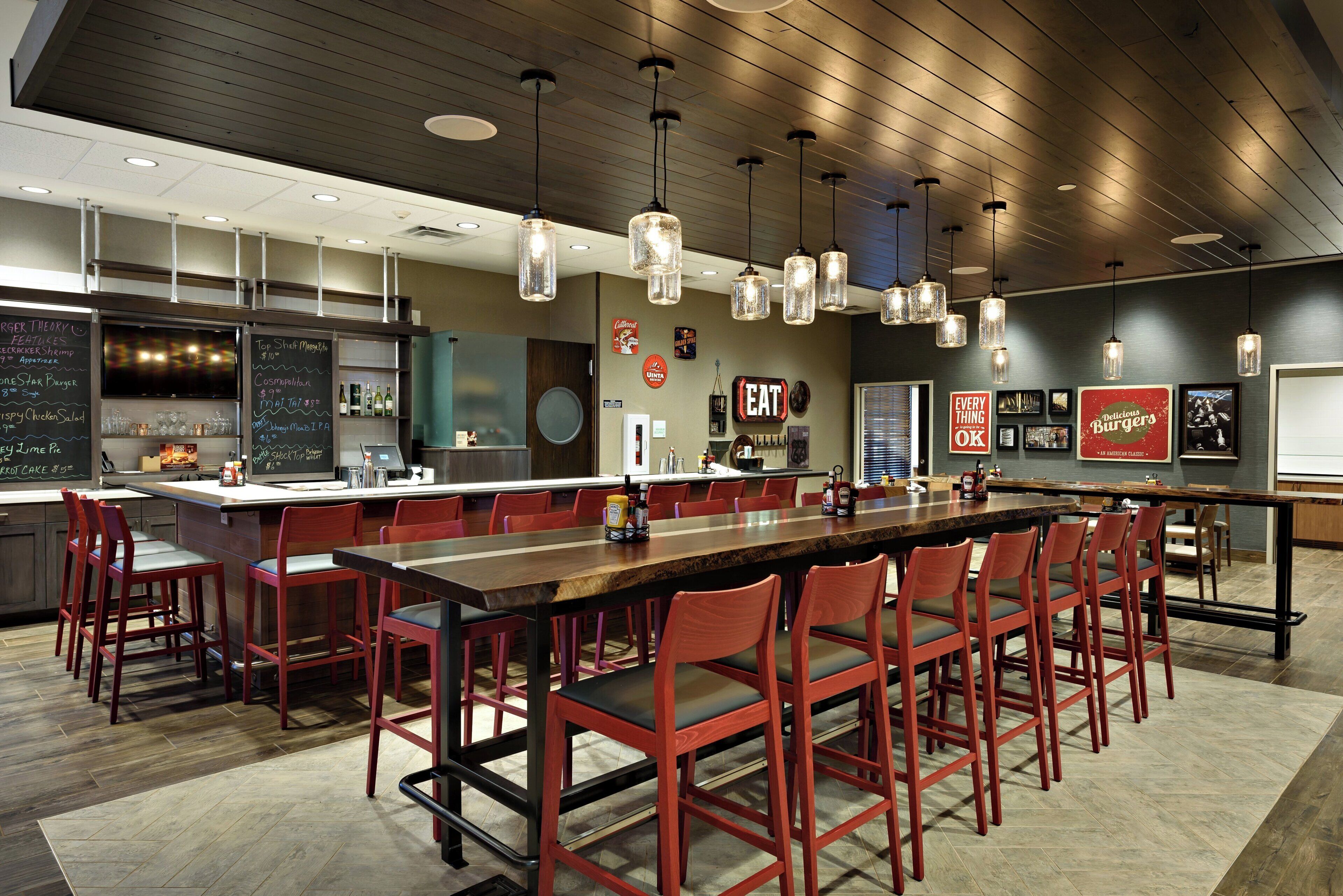 A modern restaurant with a long communal table, red chairs, pendant lighting, and a chalkboard menu.