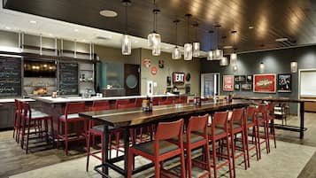 A modern restaurant with a long communal table, red chairs, pendant lighting, and a chalkboard menu.