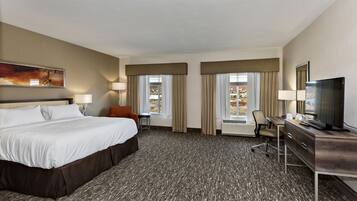 A hotel room with a large bed, a desk with a computer, a TV, and two windows with curtains.