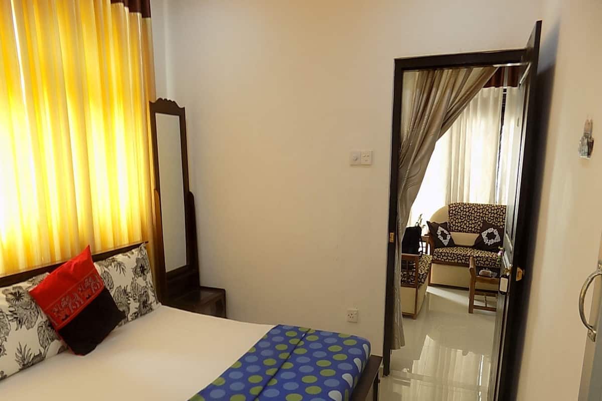 A bedroom with a bed, a nightstand, a mirror, and a view into another room with a sofa and curtains.