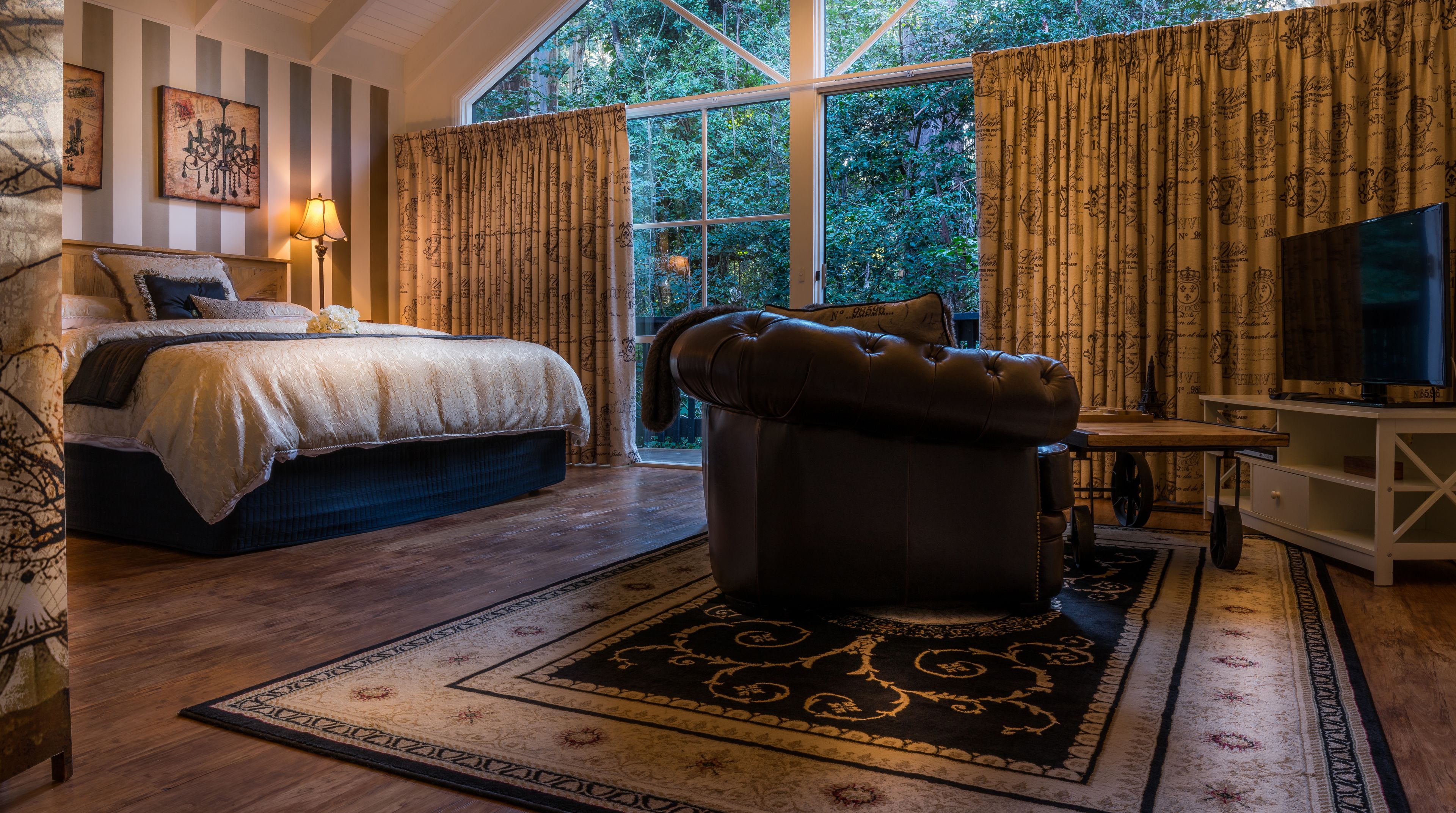 Quarto com uma cama grande, um banquinho de couro, televisão e janela com cortinas.