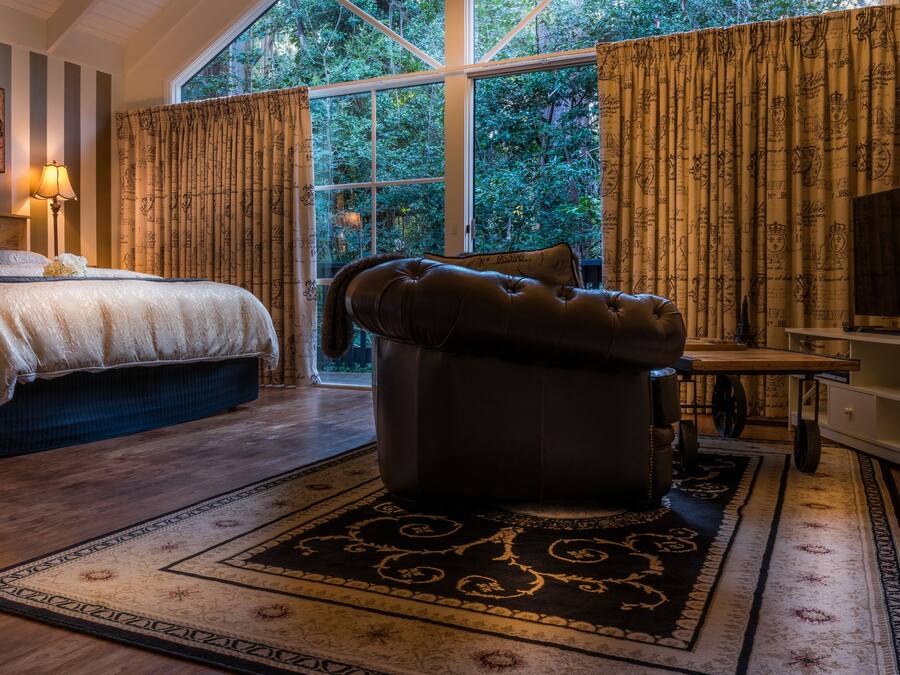 Quarto com uma cama grande, um banquinho de couro, televisão e janela com cortinas.