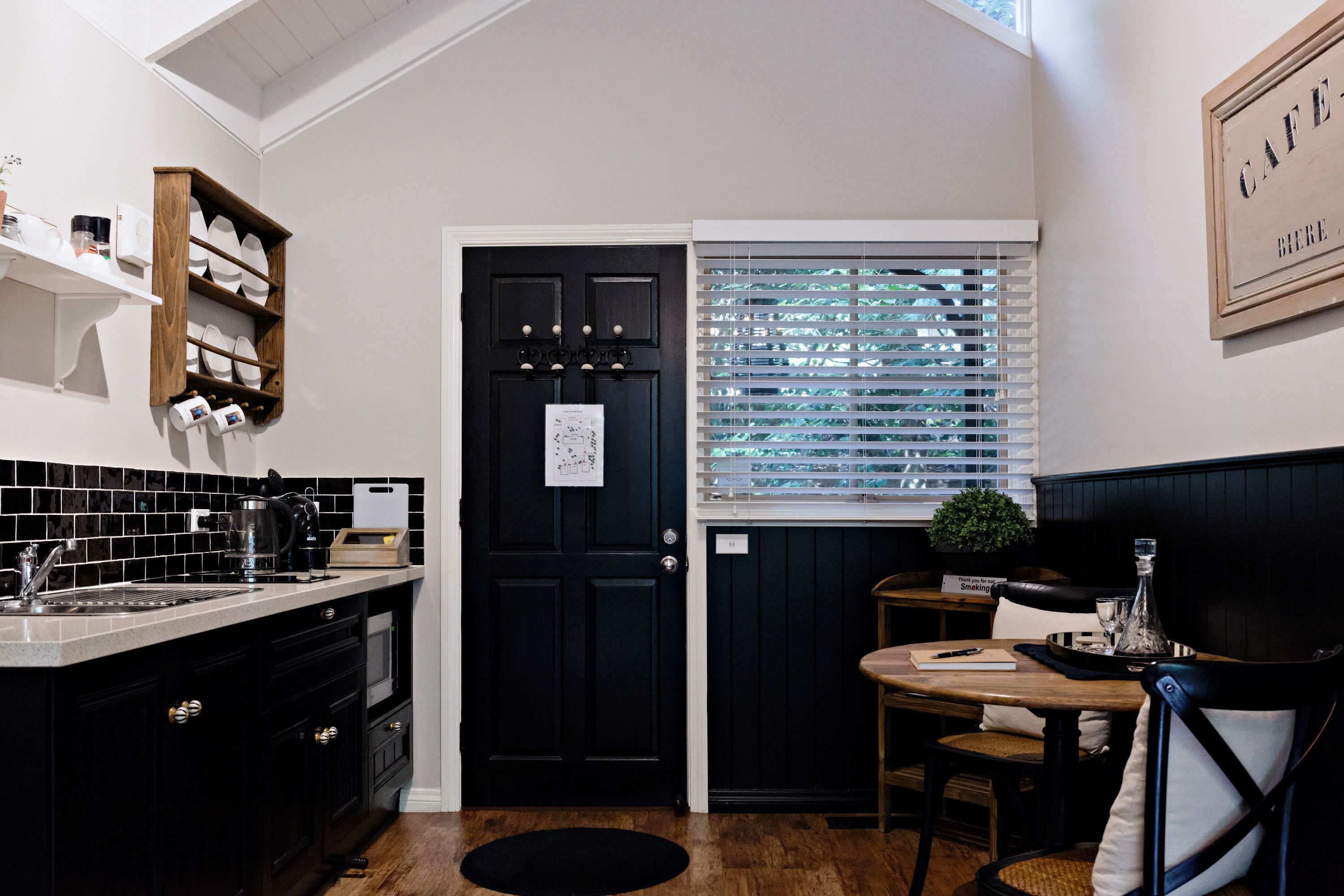 A kitchen with a black door, a window with blinds, and a dining area with a table and chairs.