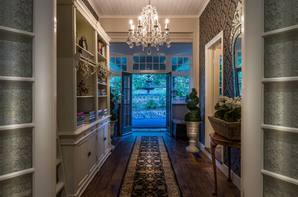 A hallway with a chandelier, open doors leading to a garden, and shelves with decorative items. - Belle Le Vie (Sassafras)