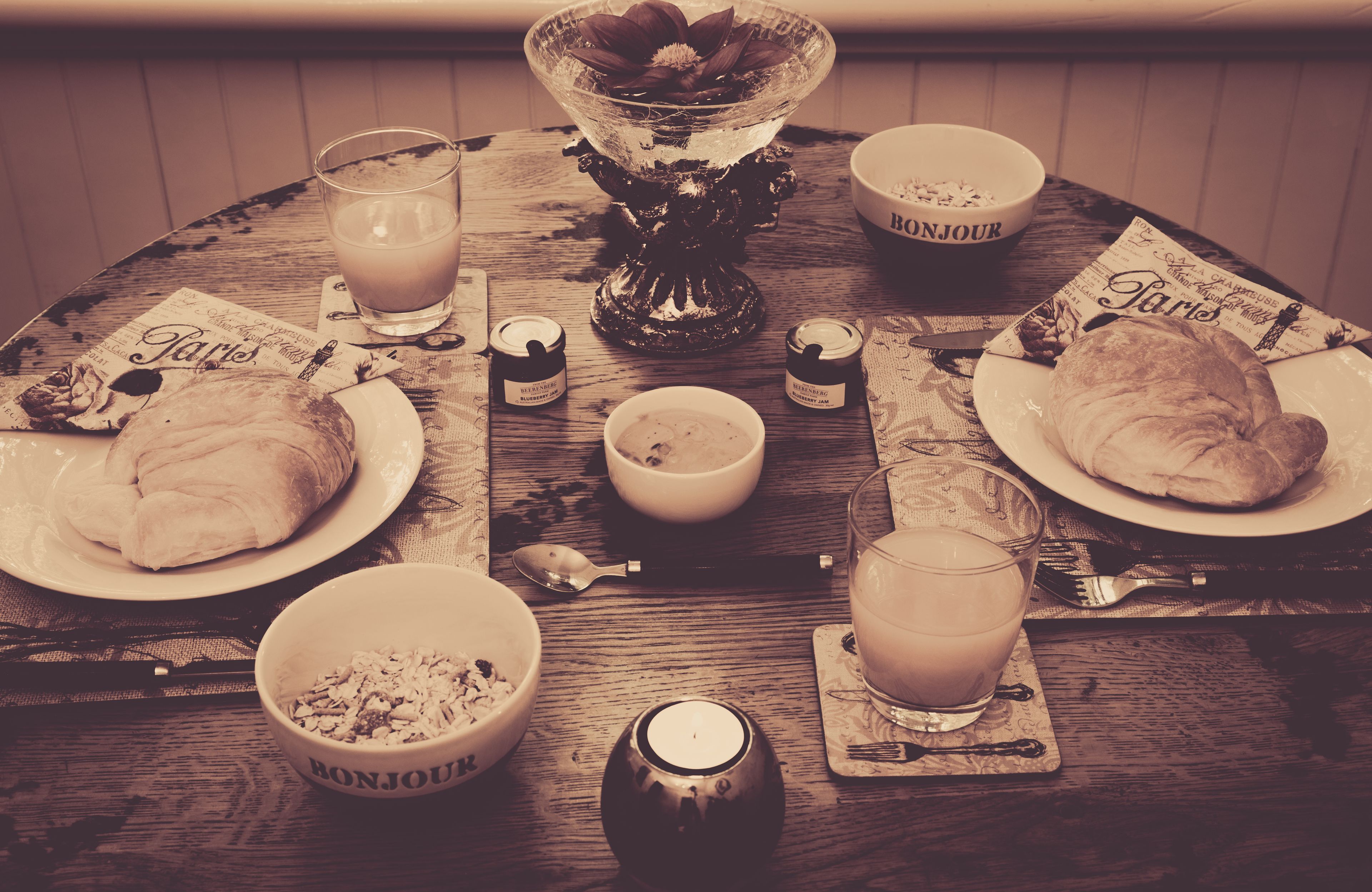 Une table ronde en bois, mise pour le petit-déjeuner, avec des croissants, un saladier de fruits, un verre de lait et une bougie.