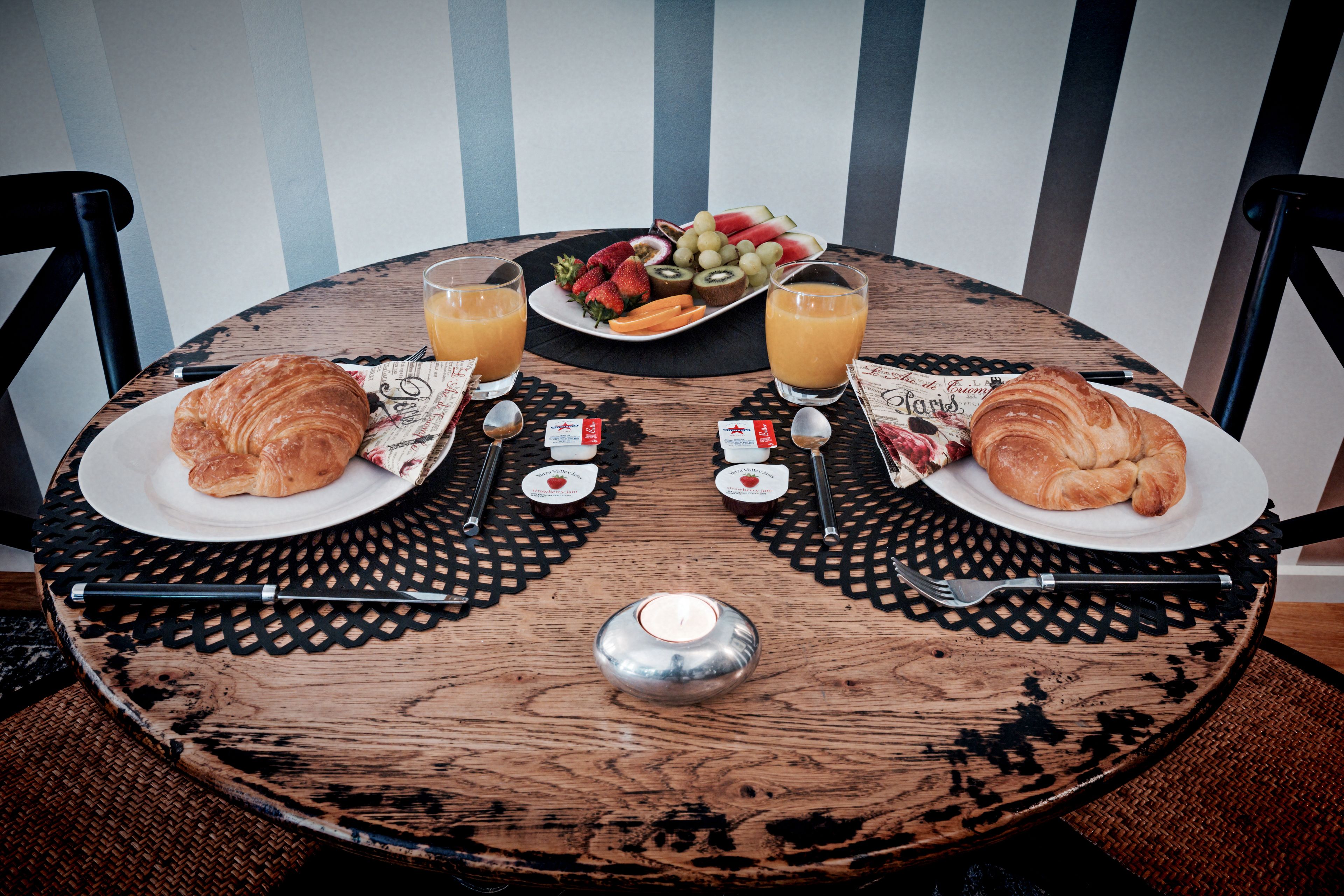 Una mesa de madera redonda con un mantel individual, dos croissants, un plato de fruta, dos vasos de zumo de naranja y una vela pequeña.