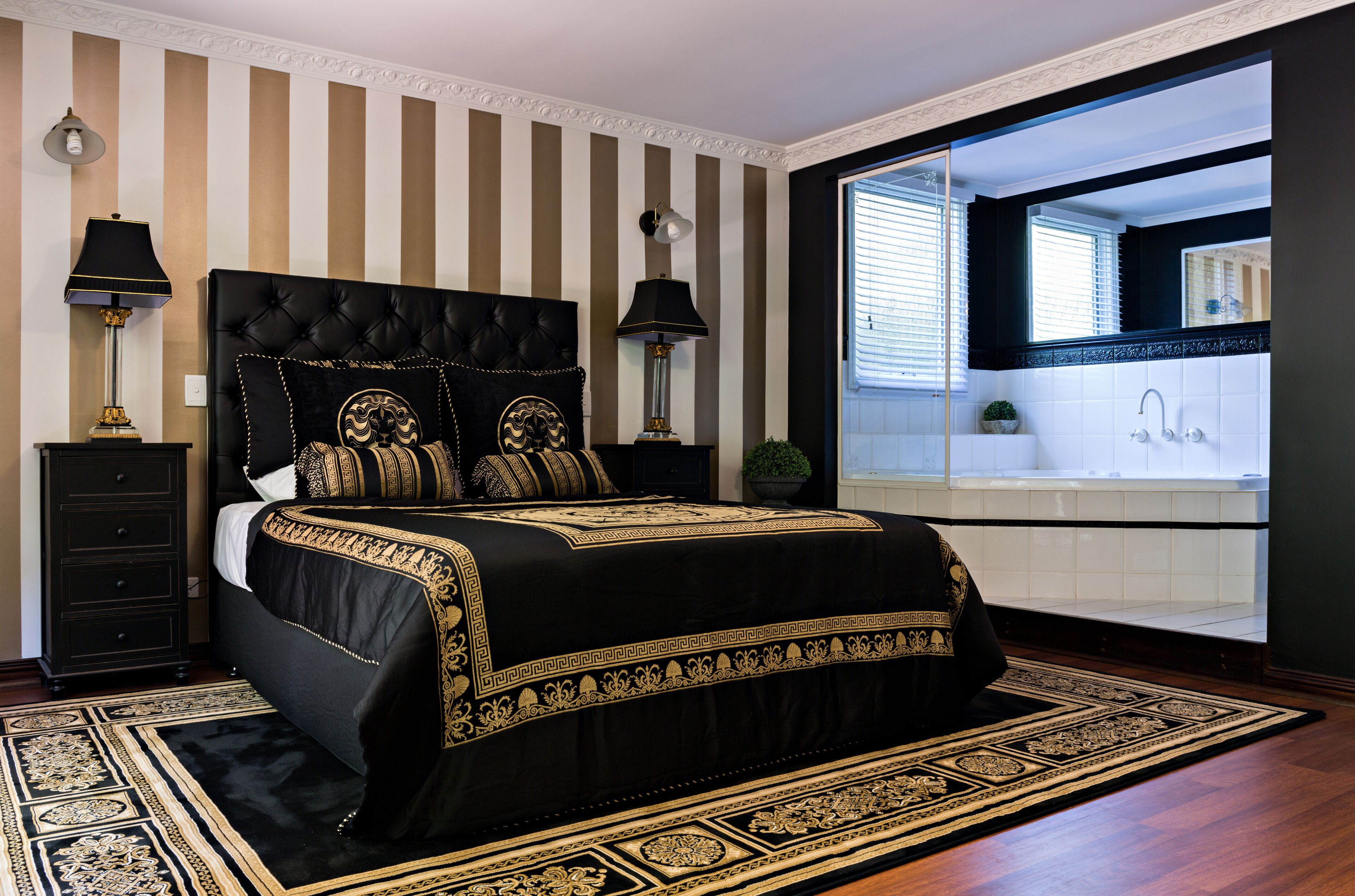 Een slaapkamer met een groot bed, twee bedlampen, een nachtkastje, een potplant en uitzicht op een badkamer met een wastafel en spiegel.