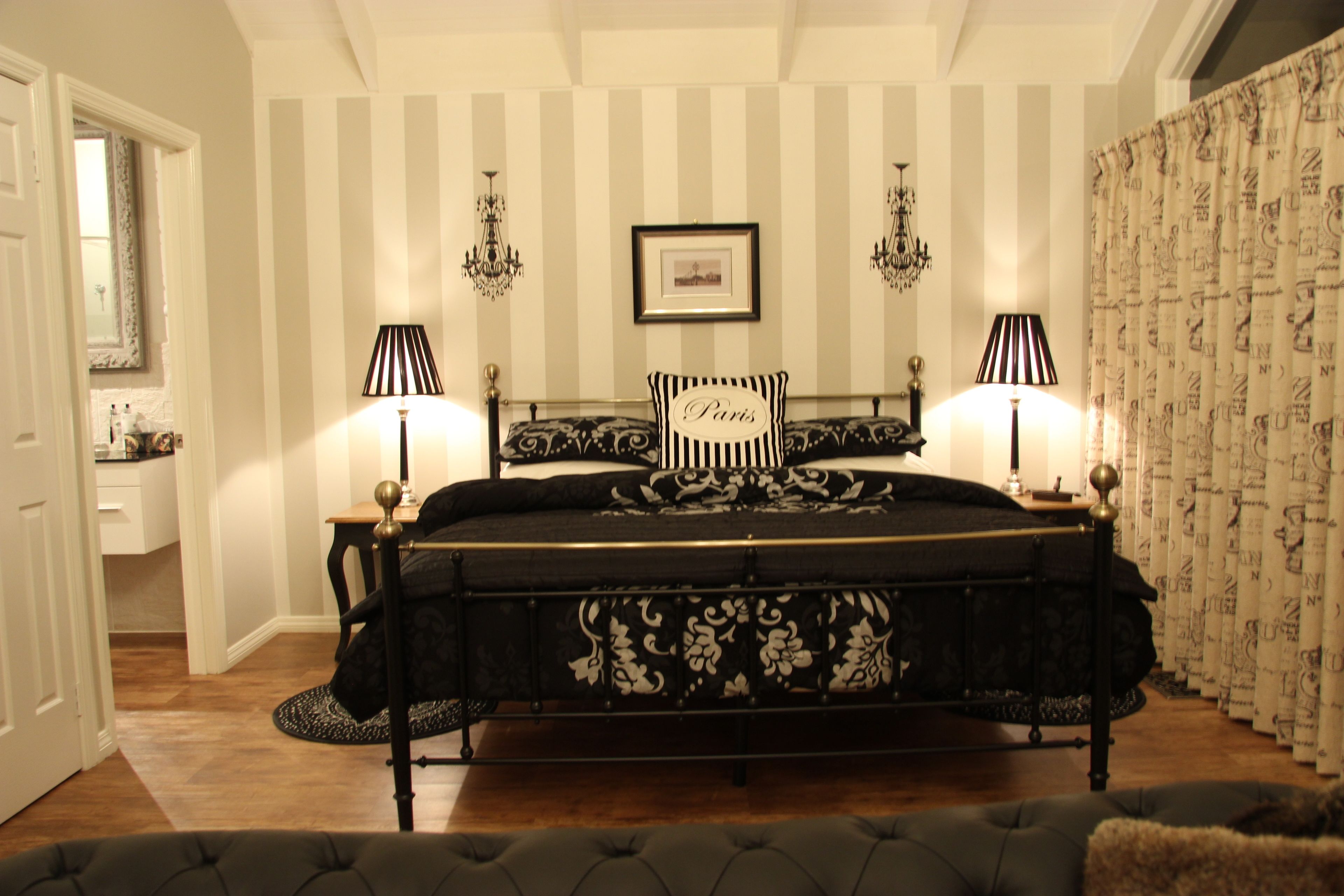 A bedroom with a bed, two bedside lamps, a chandelier, a framed picture on the wall, and striped wallpaper.