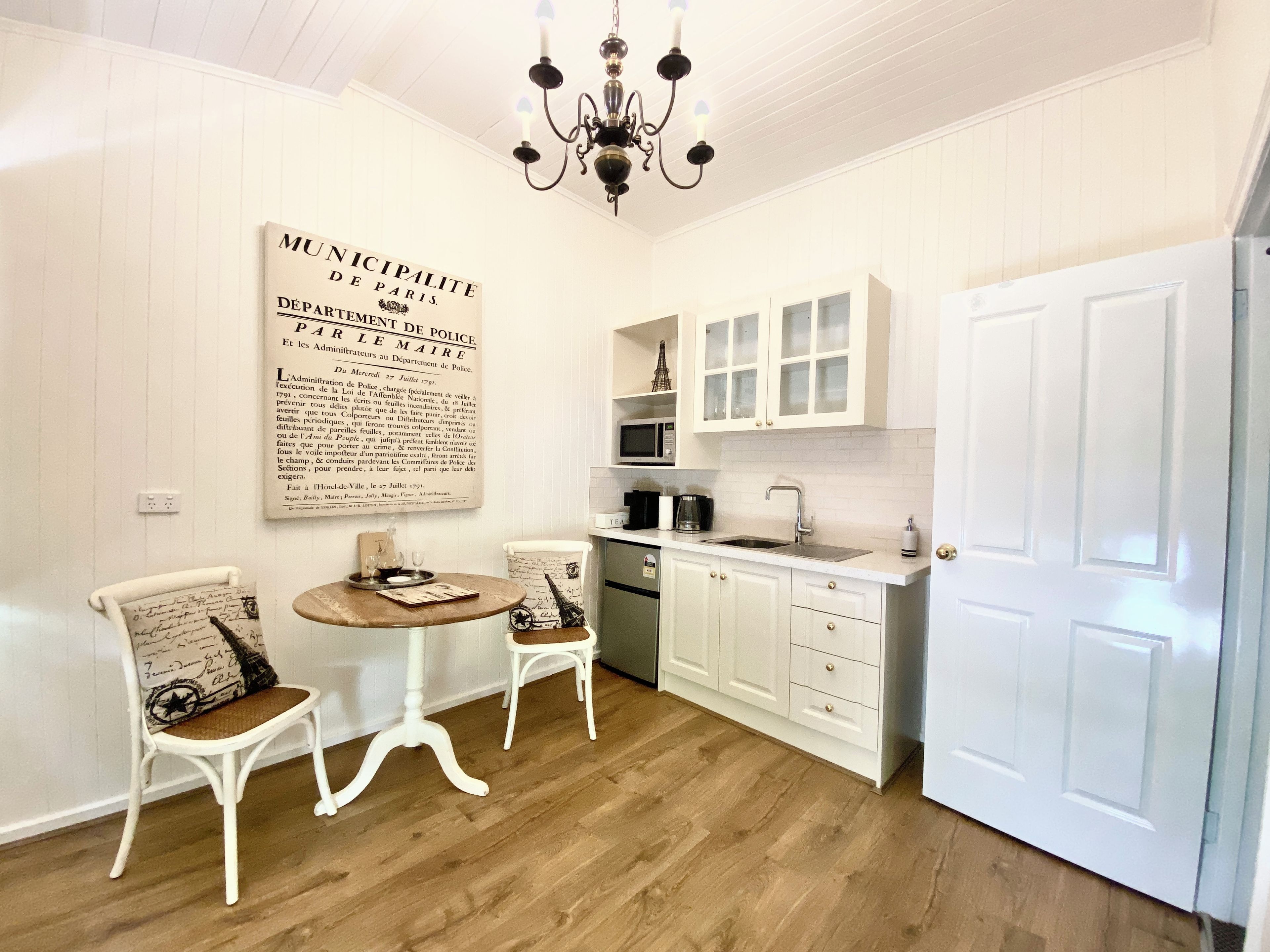 A kitchen with white cabinets, a wooden dining table, two chairs, a chandelier, and a large historical poster on the wall.