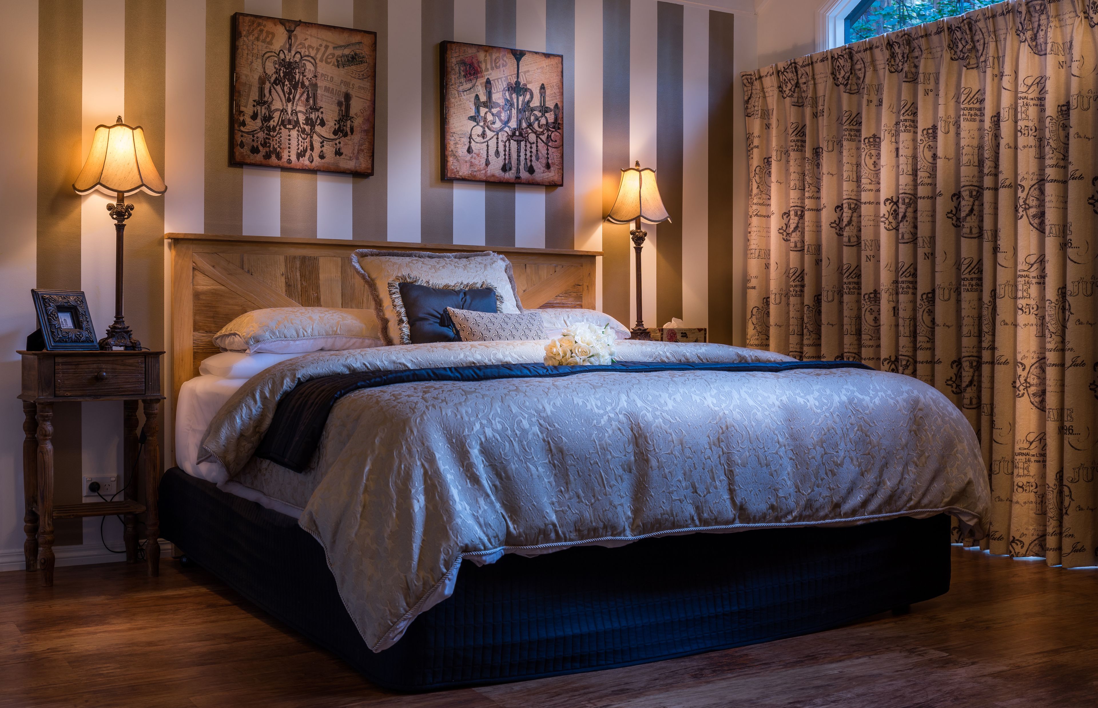 Una cama bien hecha con un cabecero de madera, dos lámparas de noche y un arreglo floral. Las paredes tienen rayas y hay una cortina con texto.