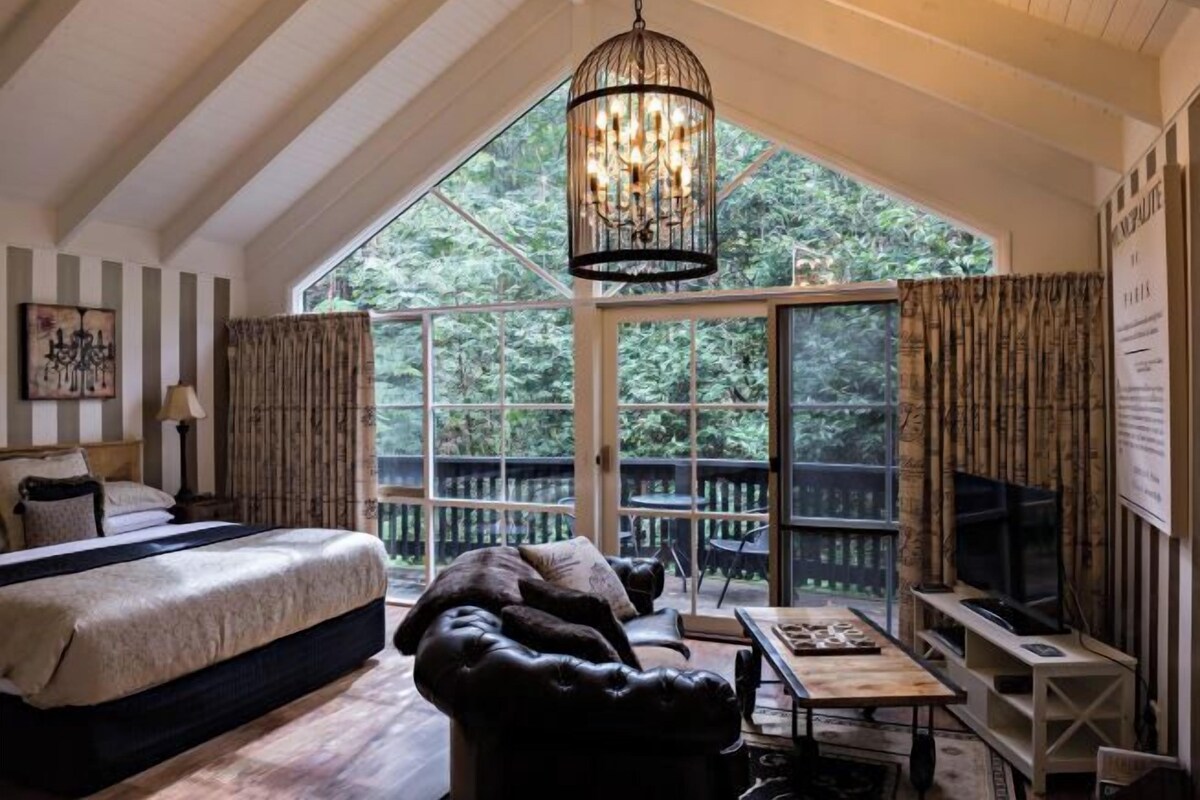 A bedroom with a large bed, a sofa, a TV, and a chandelier. There is a window with curtains and a view of trees outside.