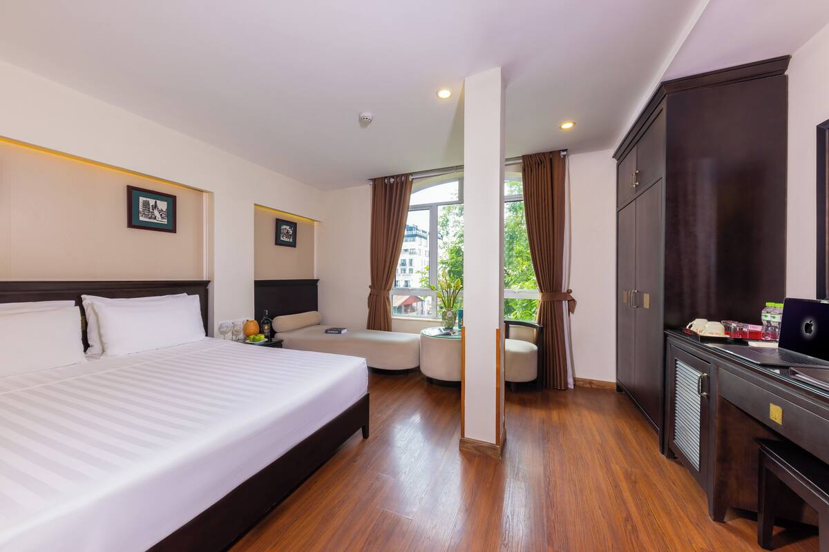 A hotel room with a large bed, a desk with a laptop, a chair, a small table with a vase, and a window with curtains.