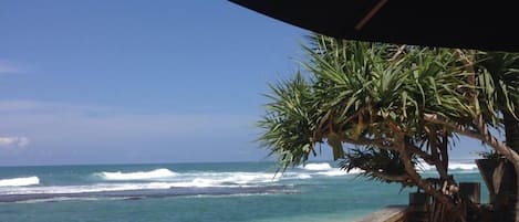 Muro de piedra que conduce a una playa con olas, una palmera y un área de descanso con cojines a rayas.