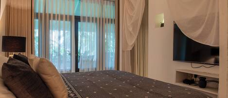 A hotel room with a bed, curtains, a television, and a shelf with items.