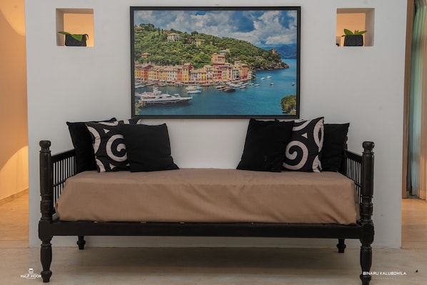 A bed with a beige cover, black frame, and two black pillows with white swirls. A framed painting of a coastal town is above the bed. - WB Villa (Unawatuna)