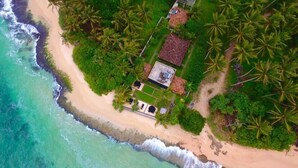 An aerial view of a coastal property with a beach, palm trees, and a small house with solar panels. - WB Villa (Unawatuna)