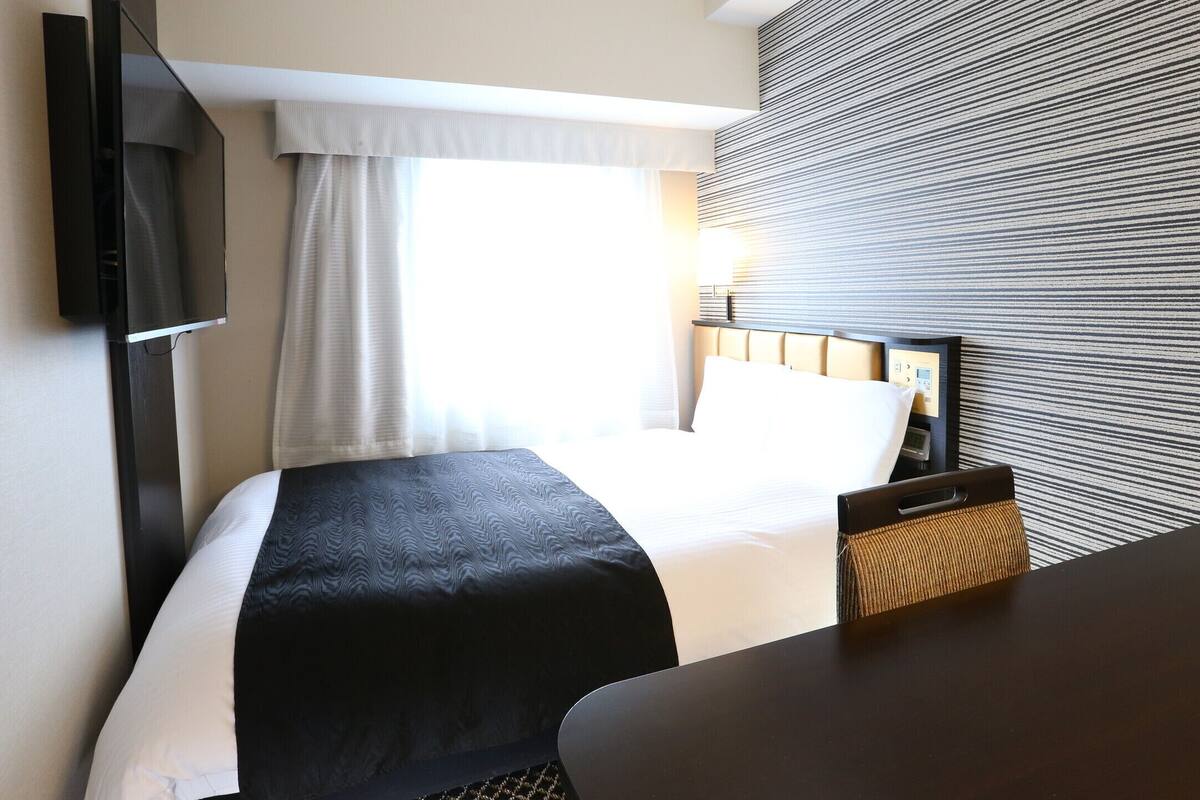 Une chambre d’hôtel avec un lit, un téléviseur fixé au mur, un bureau et une fenêtre avec des rideaux.