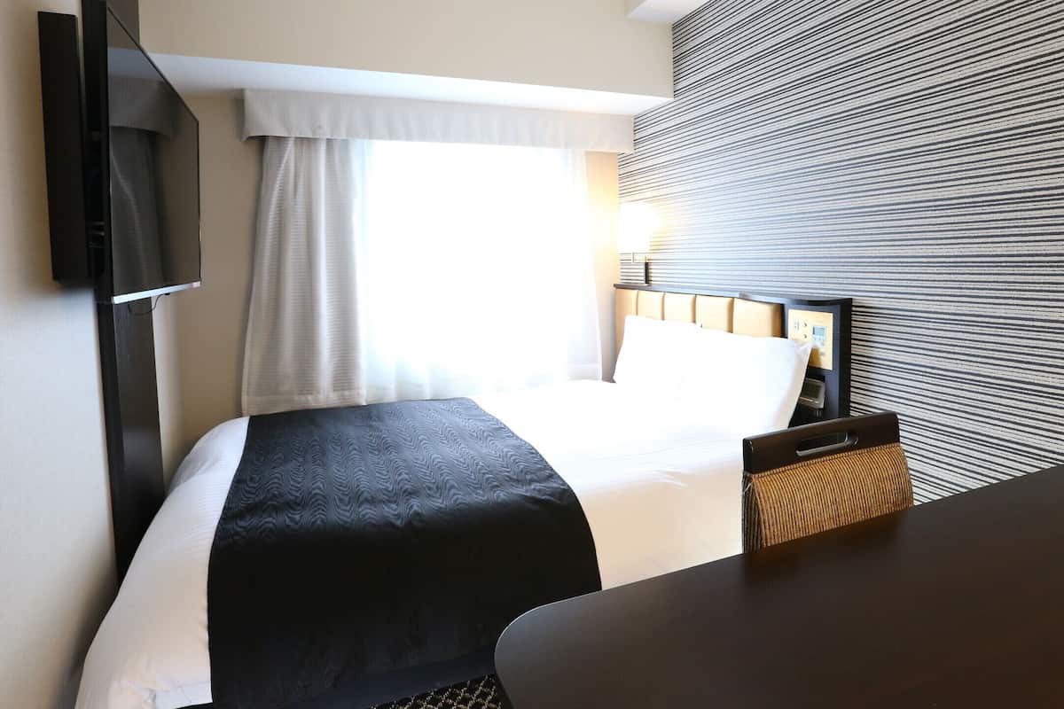 A hotel room with a bed, a TV mounted on the wall, a desk, and a window with curtains.