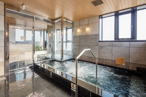 Un espace piscine intérieur moderne, doté de murs en verre, d’un plafond en bois et d’un sol carrelé.