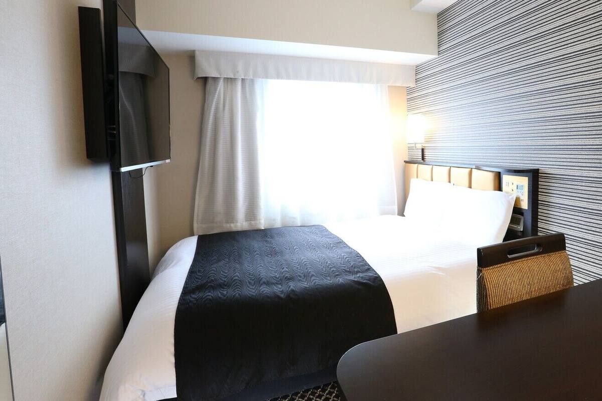 Habitación de hotel con cama, televisión montada en la pared, mesita de noche y ventana con cortinas.