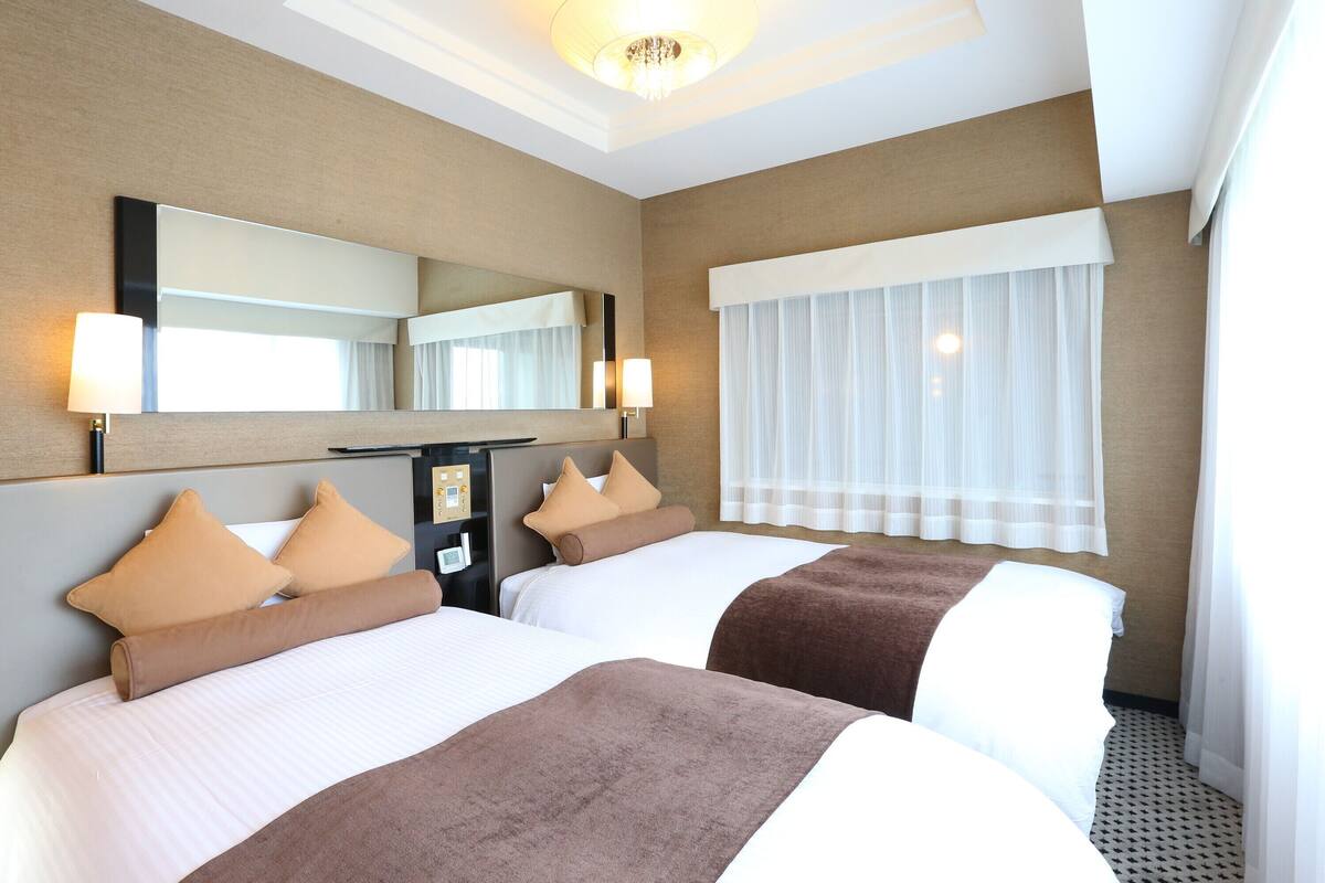 Une chambre d’hôtel avec deux lits, un grand miroir, un bureau avec une lampe et une fenêtre avec des rideaux légers.