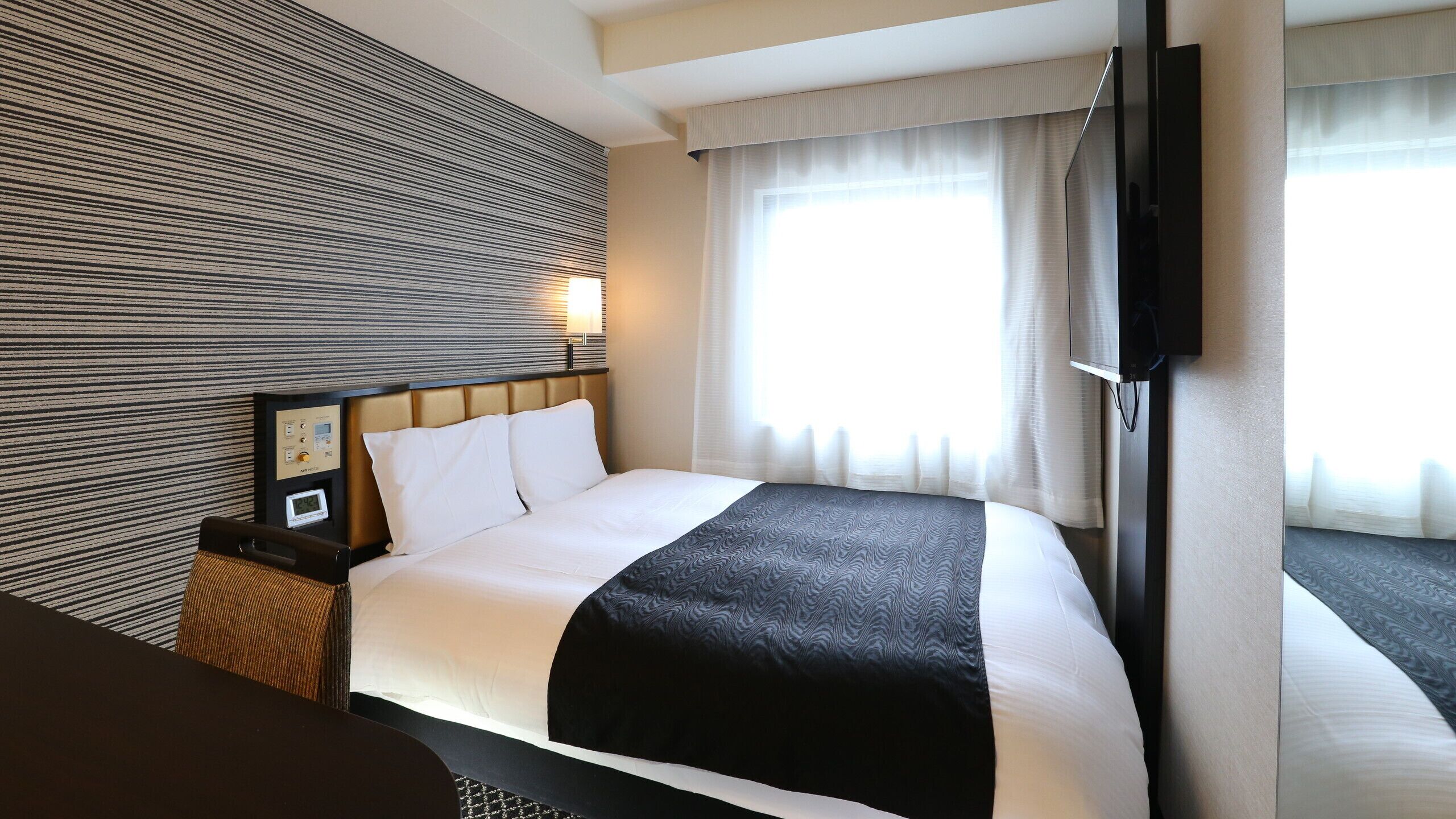 Ein Hotelzimmer mit Bett, Nachttisch mit einer Uhr, einem an der Wand montierten Fernseher und einem Fenster mit Vorhängen.