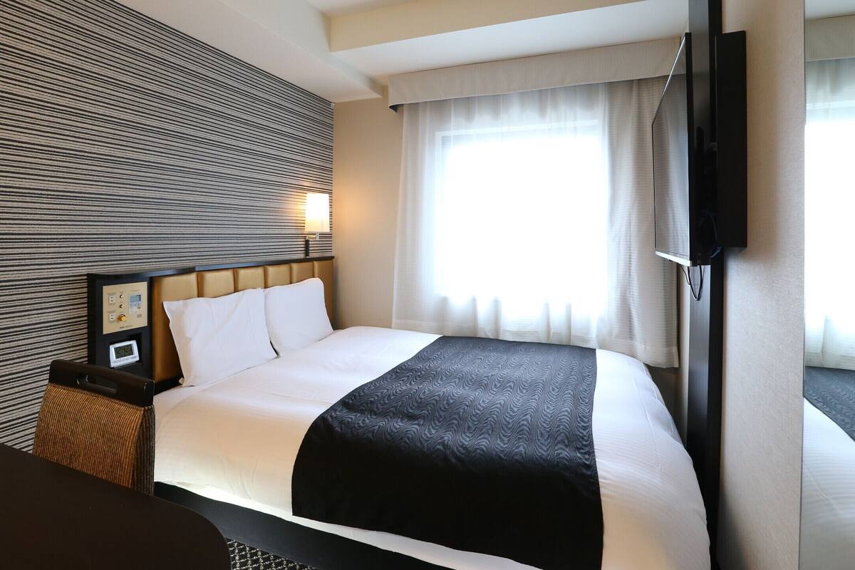 Une chambre d’hôtel avec un lit, une table de chevet avec une horloge, un téléviseur fixé au mur et une fenêtre avec des rideaux.