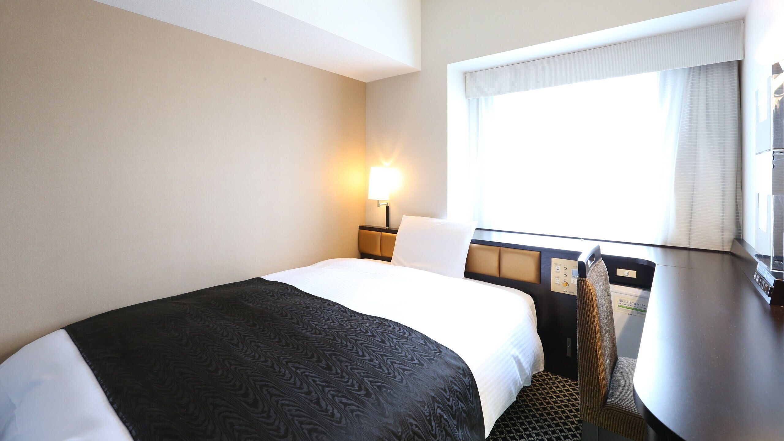 Ein Hotelzimmer mit Bett, Schreibtisch, Stuhl, einem Fenster mit Vorhängen und einer Wandlampe.