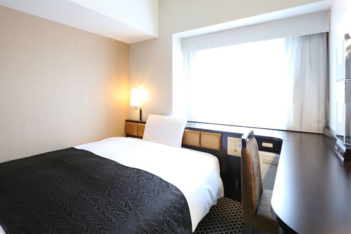 Une chambre d’hôtel équipée d’un lit, d’un bureau, d’une chaise, d’une fenêtre avec des rideaux et d’une lampe fixée au mur.